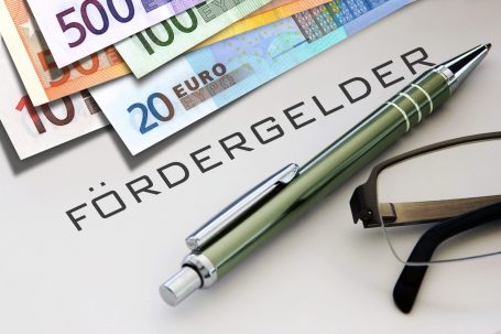 Der Hausbau Berater - Fachberater für Fertighäuser - Thomas Bärtle Eurobanknoten, ein Stift und eine Brille, mit dem Wort "Fördergelder" im Hintergrund.