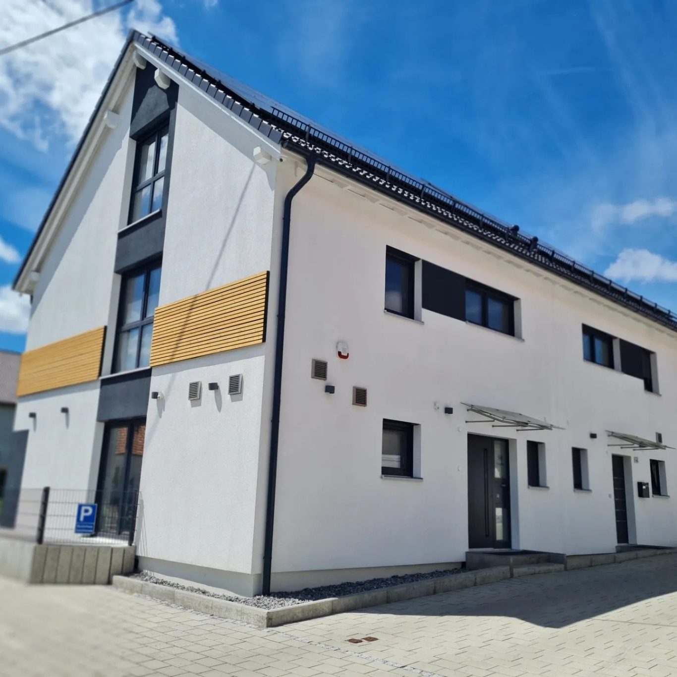 Enovia Haus / Musterhaus Altenmünster Helles modernes Gebäude mit weißer Fassade und Holzverkleidung, unter blauem Himmel.