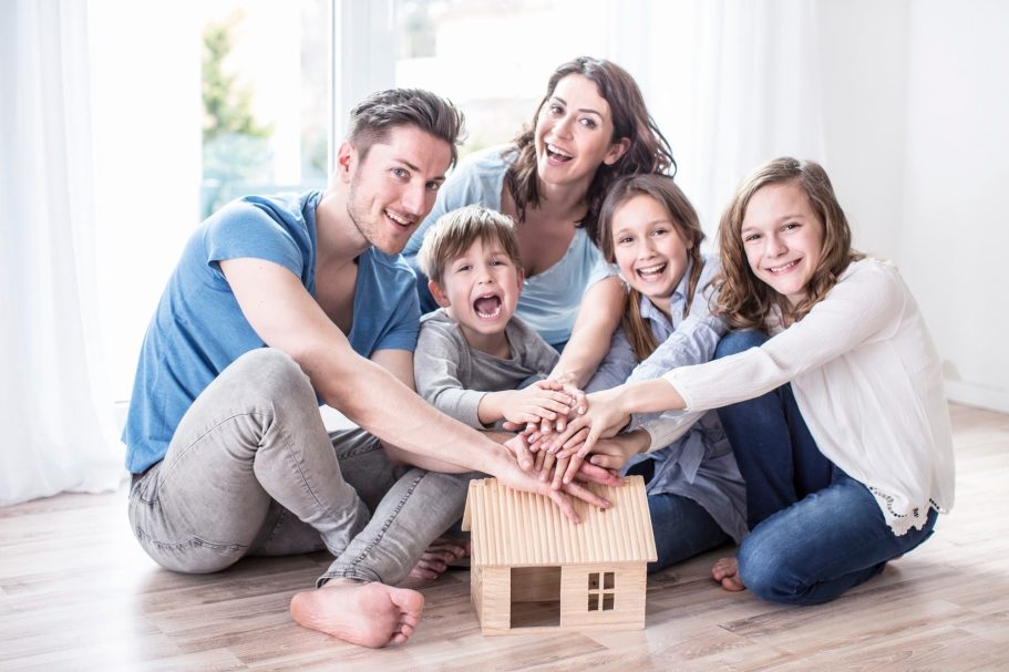 Der Hausbau Berater - Fachberater für Fertighäuser - Thomas Bärtle Eine Familie sitzt am Boden und legt ihre Hände auf ein kleines Spielhaus.